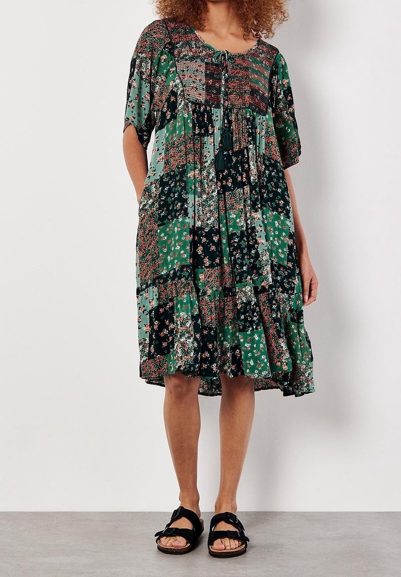 Femme portant une robe patchwork fleurie longueur genou dans des tons verts et noirs avec des manches au niveau des coudes et des sandales noires à enfiler.
