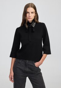 Zwarte gebreide top met een hoge kraag en een decoratieve strassaccent met een strik, evenals een combinatie met donkergrijze jeans.