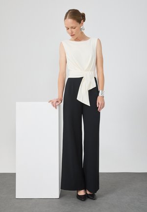 Jumpsuit mit cremefarbener Oberseite und weit geschnittenen schwarzen Hosen, mit einem vorne gebundenen Gürtel. Glatter Stoff mit minimalistischem Design.