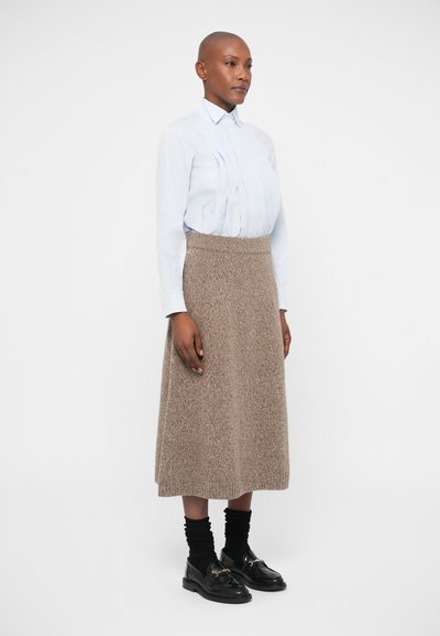 Ανοιχτό μπλε πουκάμισο με πτυχώσεις, συνδυασμένο με καφέ υφασμάτινη φούστα μέχρι το γόνατο, μαύρες κάλτσες και μαύρα loafers με ασημί λεπτομέρειες.