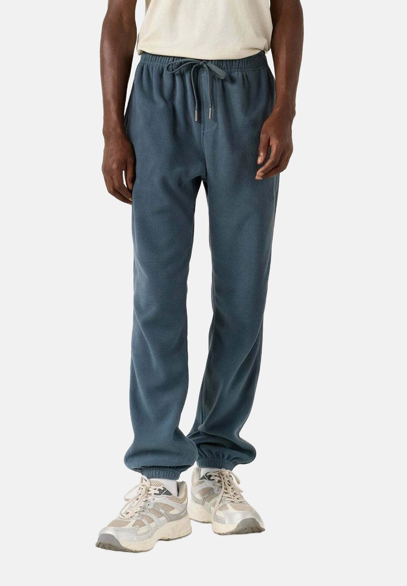 Man in donkerblauwe fleece joggingbroek en lichtbeige hardloopschoenen, staand tegen een effen lichte achtergrond.