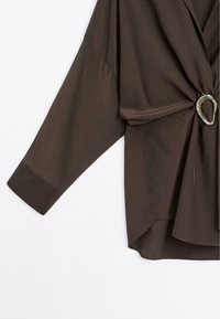 Blouse marron foncé à manches longues avec une large manchette et un détail de nœud torsadé à l’avant maintenu par une boucle anneau argentée.
