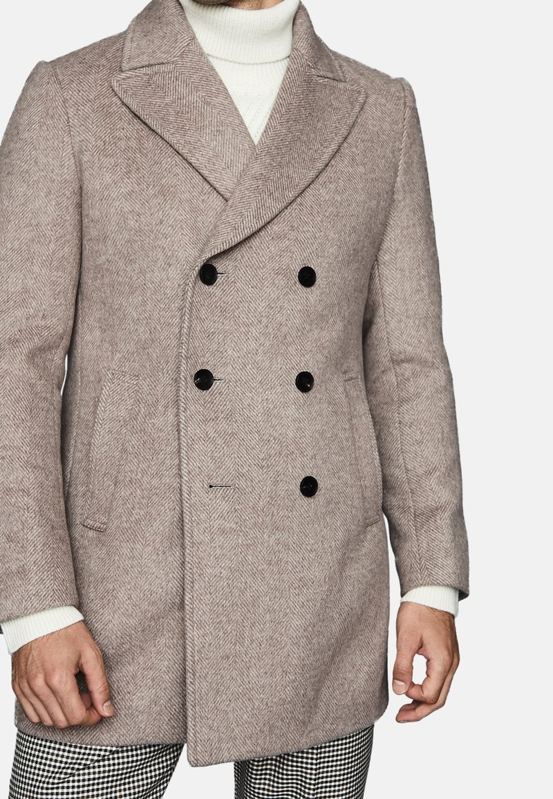 Cappotto doppiopetto in leggero beige a motivo herringbone, con bottoni neri, colletto a scollatura a V e tasche laterali. Maglione bianco a maglia sotto.