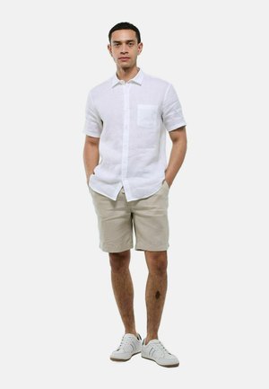 Chemise en lin blanc à manches courtes avec une poche poitrine, associée à un short beige et des baskets blanches, se tenant les mains dans les poches.