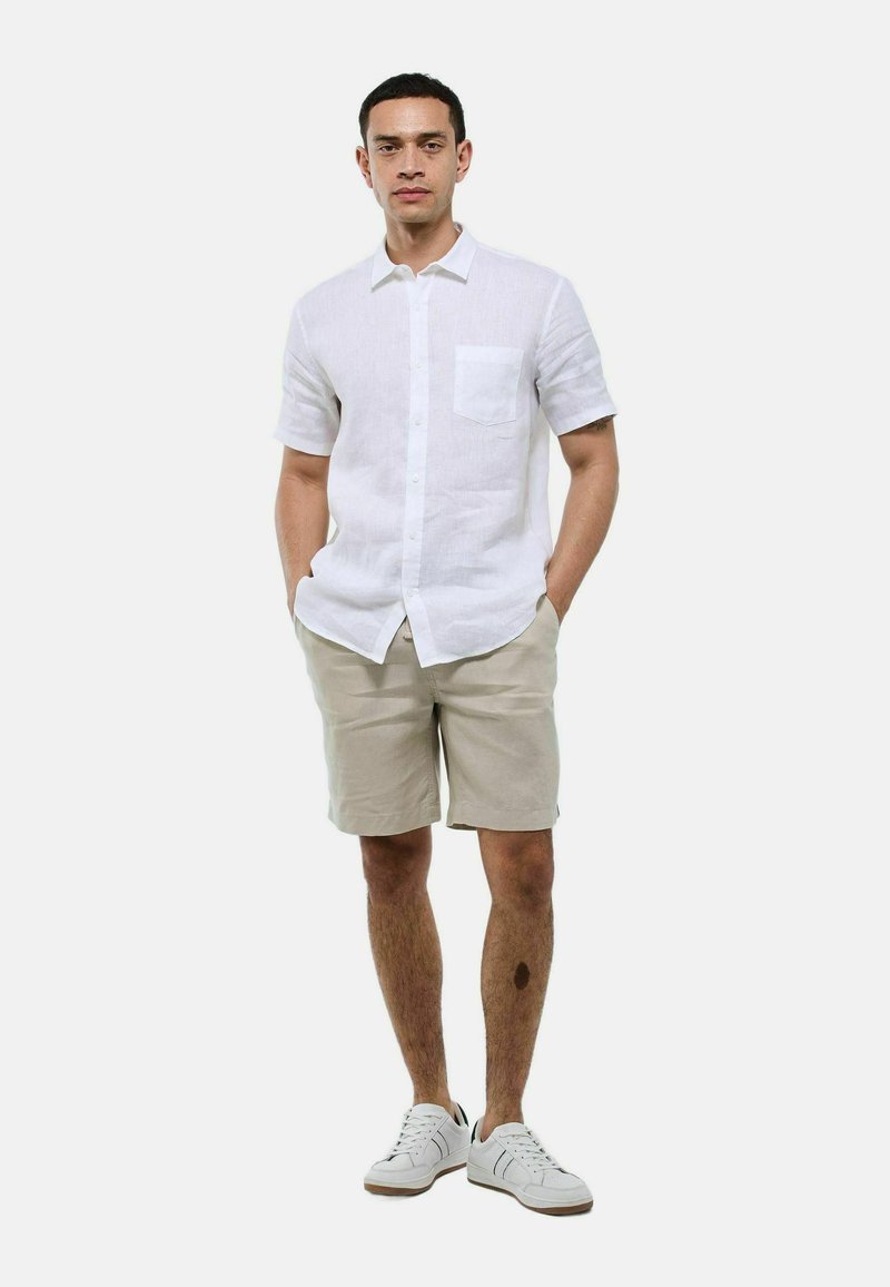Chemise en lin blanc à manches courtes avec une poche poitrine, associée à un short beige et des baskets blanches, se tenant les mains dans les poches.