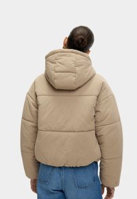 Beige polstret jakke med en stor hætte, der har et segmenteret design, blød tekstur og elastisk kant. Båret over lys blå jeans.