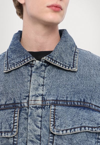 Jachetă din denim de culoare albastru deschis, cu o finisare texturată, având un guler clasic, două buzunare pe piept și cusături contrastante.