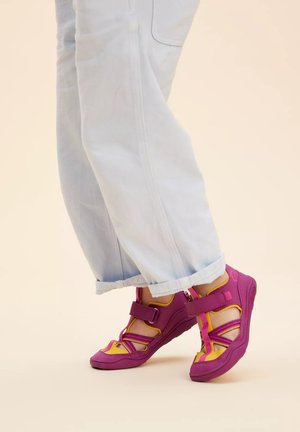Lila und gelbe Sandalen mit verstellbaren Riemen, robusten Gummisohlen und durchbrochenem Design, getragen mit hellblauen, umgeschlagenen Jeans.