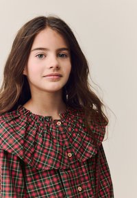 Blouse à carreaux rouges et verts avec un col à volants, tissu froncé et boutons sur le devant, fabriquée en mélange de coton doux.