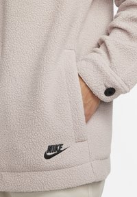 Fleece-pulloverjakke i lyserosa, med en teksturert overflate, sidelommer og svart Nike-logo nær kanten. Knappe detaljer ved mansjetten.