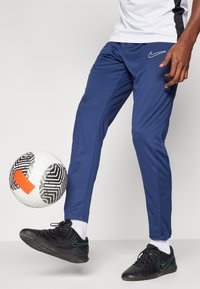 Jūras spēku sporta bikses ar šauru piegriezumu, uz augšstilba ir Nike logo, savienotas ar melniem sporta apaviem un rakstainu futbola bumbu.