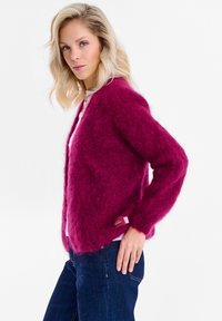 Blondynka ubrana w puszysty, magentowy kardigan na jasnoróżowym topie i ciemnoniebieskich dżinsach, pozująca bokiem z ręką na biodrze.