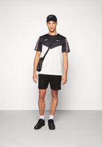 Camiseta Nike en blanco y negro con un diseño de chevron, pantalones cortos negros y una gorra negra. El modelo lleva una bolsa de hombro negra.