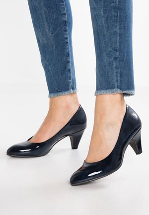 Kvinde iført mørkeblå laklæder midt-hælspumper med slidte blå jeans, stående på et hvidt gulv.