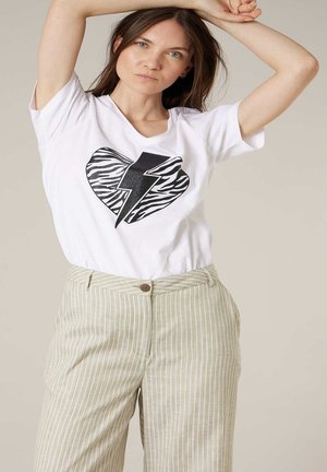 Vrouw draagt een wit T-shirt met een hart en bliksemschicht in zebraprint, gecombineerd met beige gestreepte broek, poseert met opgeheven armen.