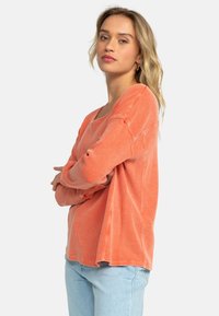 Pitkähihainen, korallinoranssi toppi ribbineuloksesta, jossa on lasketut olkapäät ja sivuhalkiot. Yhdistetty vaaleansinisiin farkkuihin.