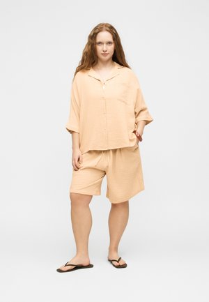 Kvinde med langt krøllet hår iført løs beige skjorte med knapper og matchende shorts, stående barfodet i sorte flip-flops på ensfarvet baggrund.