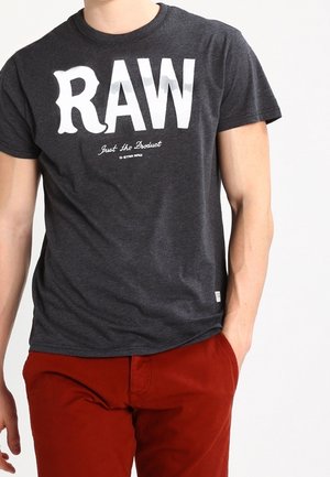 Mężczyzna w ciemnoszarej koszulce z dużym białym napisem „RAW” i czerwonych spodniach, stojący z jedną ręką w kieszeni na białym tle.