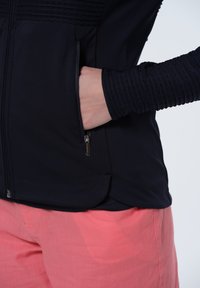 Svart zip-up väst med texturerad axeldetalj, sidaficka och slät tyg. I kombination med ljusrosa byxor.