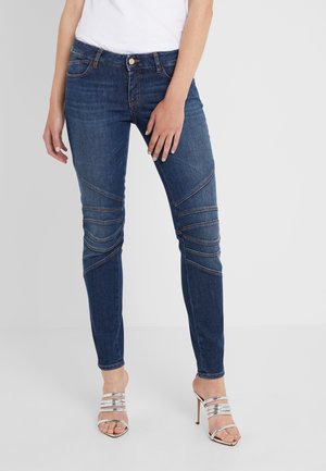 Nainen, joka pukeutunut tummansinisiin tiukkoihin farkkuihin, joissa on tikatut polvityynyt, sekä hopeisiin korkokenkiin, seisoo yksinkertaisen vaalean taustan edessä.