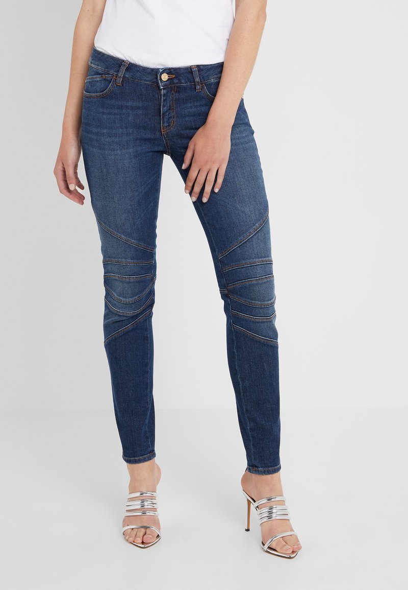 Femme portant un jean skinny bleu foncé avec des détails de couture au niveau des genoux et des sandales à talons hauts argentées, debout devant un fond clair uni.