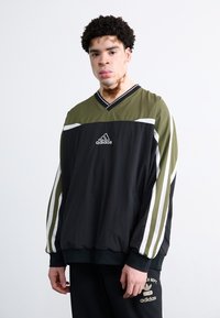 Svart och olivgrön jacka med V-ringning, vita ränder längs ärmarna och en broderad Adidas-logotyp på bröstet. Har en lätt texture.