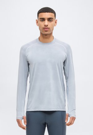 Hombre con camiseta deportiva de manga larga gris claro y mallas deportivas gris oscuro, de pie mirando hacia adelante sobre un fondo liso.