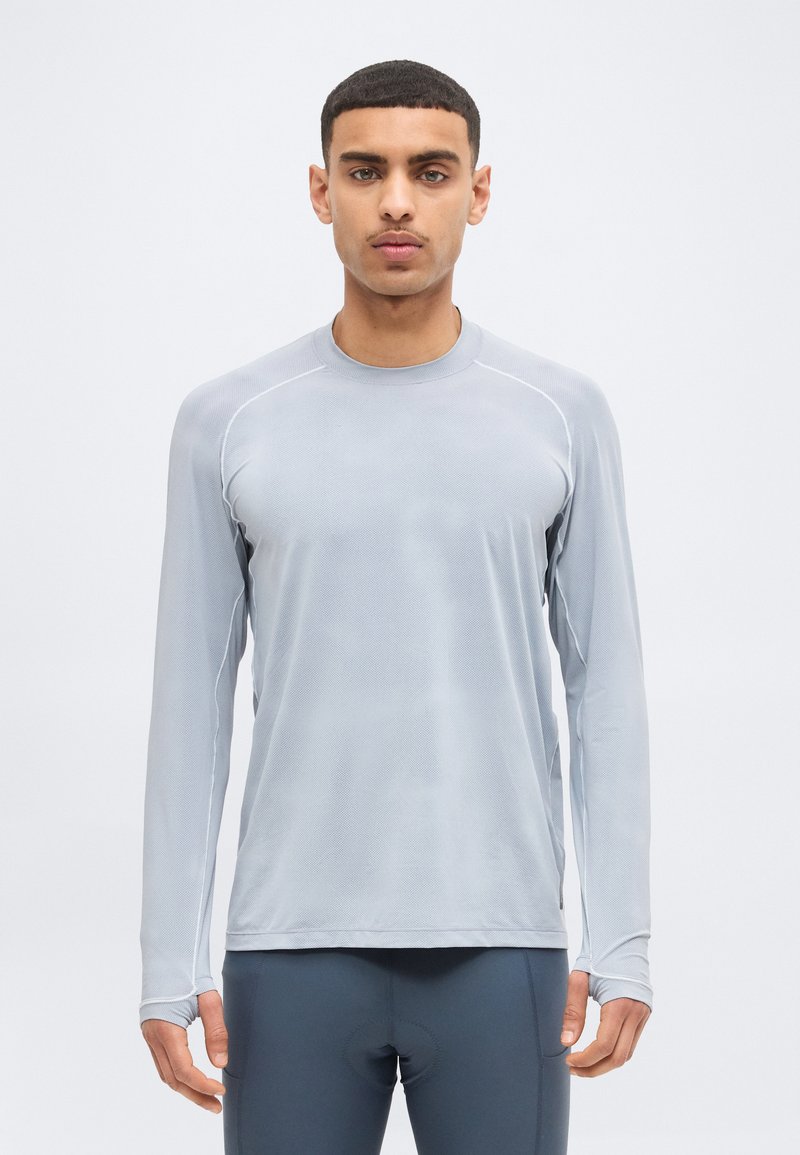 Άνδρας που φοράει ανοιχτό γκρι αθλητικό πουκάμισο με μακριά μανίκια και σκούρο γκρι αθλητικό κολάν, στέκεται με το πρόσωπο προς τα εμπρός μπροστά σε απλό φόντο.