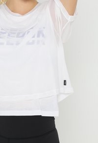 Vit nätoberdel med kort ärm och lös passform, med ett subtilt grafiskt mönster, lagerdesign och en liten logotags vid fållen.