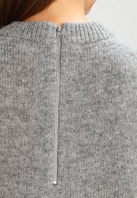 Šedý pletený svetr s vysokým žebrovaným výstřihem a viditelným podložním zipem vzadu. Měkká textura s jemným melírovaným vzorem.