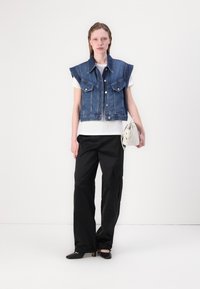 Sininen denim-leikattu liivi, jossa on olkapään läpät, kaksi etutaskua, yhdistetty valkoisen T-paidan ja mustien leveiden housujen kanssa, pitäen valkoista laukkua.