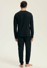 Man stands barefoot wearing a black long-sleeve top and matching black jogger pants, facing away against a plain light background.