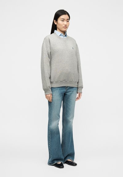 Γυναίκα που στέκεται φορώντας ανοιχτογκρι φούτερ πάνω από μπλε πουκάμισο με γιακά, καμπάνα μπλε τζιν και μαύρα παπούτσια με μυτερή μύτη, με φόντο λευκό.