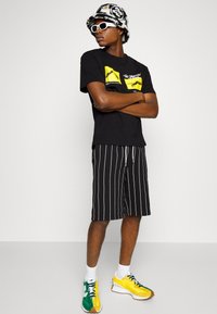 Camiseta gráfica negra con diseño amarillo, pantalones cortos a rayas en blanco y negro, gafas de sol blancas y zapatillas deportivas multicolores. Incluye sombrero tipo bucket.