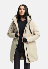 Vrouw in een beige gewatteerde winterjas met een capuchon over een zwarte top en zwarte spijkerbroek, bezig met het aanpassen van de rits van de jas, staande tegen een witte achtergrond.