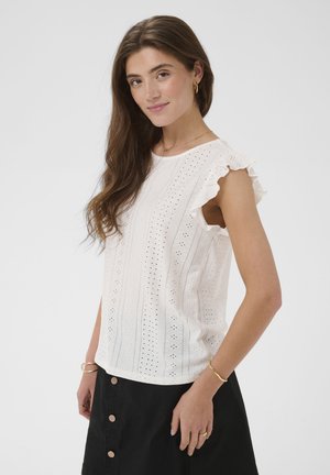 Femme aux longs cheveux bruns portant un haut sans manches en broderie anglaise blanche avec des épaules à volants et une jupe noire boutonnée, posant devant un fond blanc.