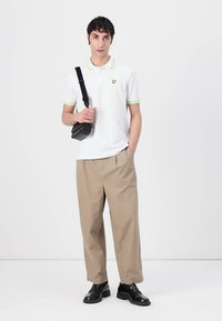 Polo bianco con accenti verdi, pantaloni beige e scarpe nere. Il modello tiene una borsa a tracolla nera, in piedi contro uno sfondo neutro.