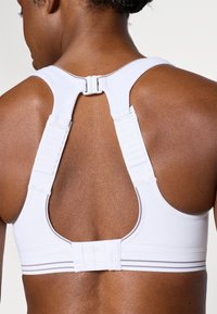 Vitt sport-bh med breda axelband, racerback-design, justerbara hakar och grå accentlinjer. Materialet verkar slätt och stretchigt.