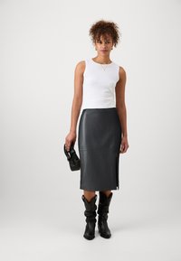 Jupe crayon en cuir noir avec une fente sur le côté, associée à un haut sans manches blanc et des bottes de cowboy noires. Tient un petit sac à main noir.