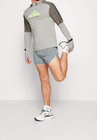 Hombre con sudadera con capucha Nike Trail gris, pantalones cortos grises y zapatillas blancas estirando la pierna derecha mientras se apoya en la pierna izquierda contra un fondo liso.