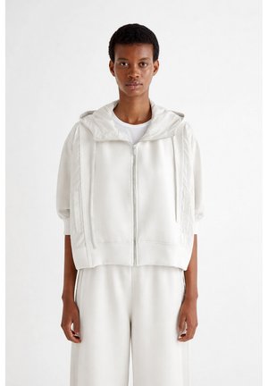 Osoba ubrana w luźną białą bluzę z kapturem na zamek i krótkimi rękawami oraz pasujące szerokie białe spodnie, stojąca na jednolitym tle.