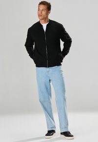 Giacca bomber nera con zip e polsini a coste, indossata sopra una camicia bianca. I jeans dritti azzurri e le sneakers nere completano l'outfit.