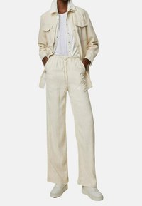 Jasnobeżowa lniana kurtka koszulowa z dwiema kieszeniami na piersi, w połączeniu z pasującymi luźnymi spodniami i białymi trampkami. Klasyczny, casualowy design.