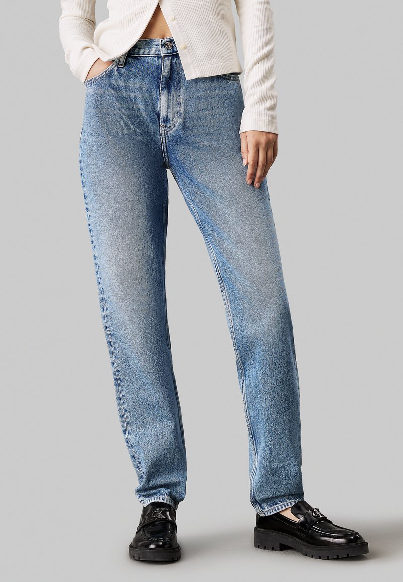 Vaqueros de mezclilla azul claro con un ajuste relajado y un acabado desgastado; cuentan con cinco bolsillos, trabillas para cinturón y un diseño de pierna recta. Combinados con mocasines negros.