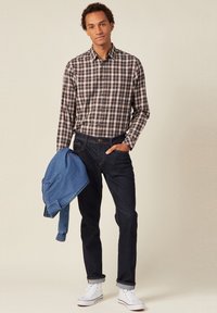 Gevlekt shirt in bruin en marineblauw, donkere spijkerbroek, houdt een blauwe spijkerjack vast, witte sneakers, staat met één hand in de zak.