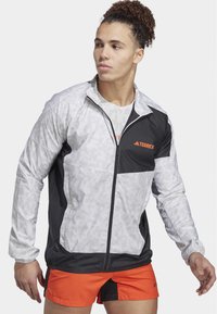 Leichte Terrex-Jacke mit einem schwarz-weißen Streifenmuster und orangenen Akzenten, durchgehendem Reißverschluss und sportlicher Passform. Kombiniert mit orangefarbenen Shorts.
