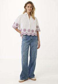 Blusa bianca in lino con orlo floreale ricamato in viola e maniche larghe, abbinata a jeans di denim a vita alta blu chiaro e sneakers beige.