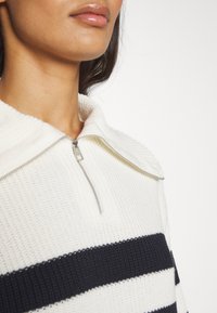 Femme portant un pull en maille blanc avec des rayures horizontales noires et une fermeture éclair argentée près du col sur un fond uni.