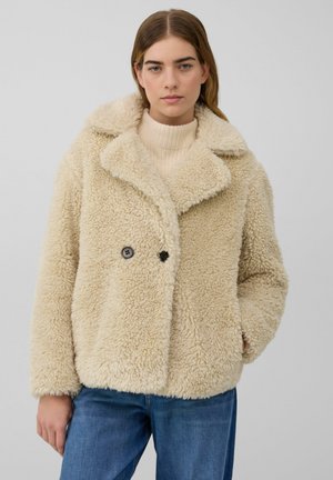 Femme portant une veste en shearling beige duveteuse par-dessus un pull en tricot crème et un jean bleu, se tenant contre un fond clair uni.