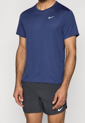 Homme portant un t-shirt Nike bleu marine à manches courtes et un short de sport Nike noir, debout devant un fond uni.